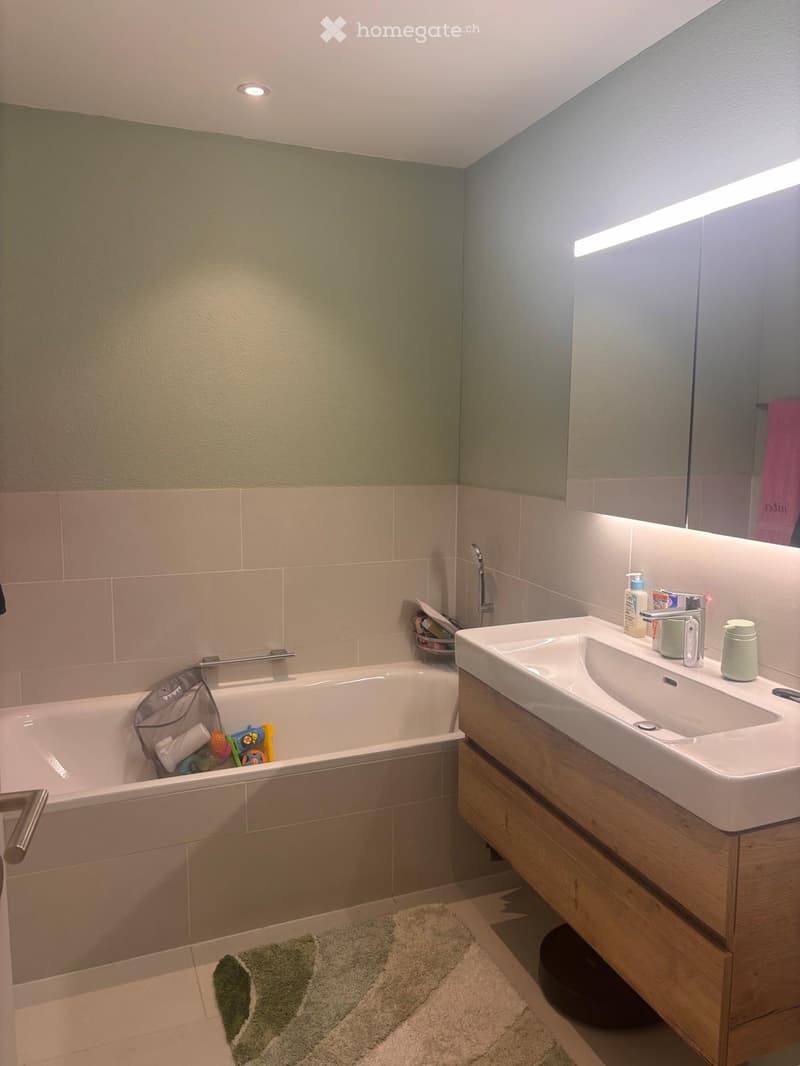 Modern bathroom with a bathtub, double sink, and a large mirror. The walls are painted in a two-tone design with green upper half and beige lower half. The floor has beige tiles with a green and beige bath mat in front of the sink.