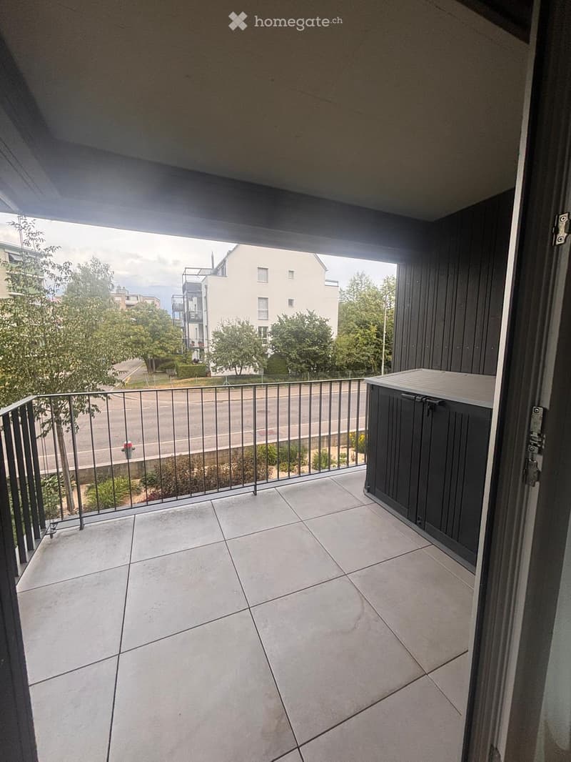Balcony with tiled floor, metal railing, and a small storage unit on the right side. The view shows a street with trees and buildings in the background.