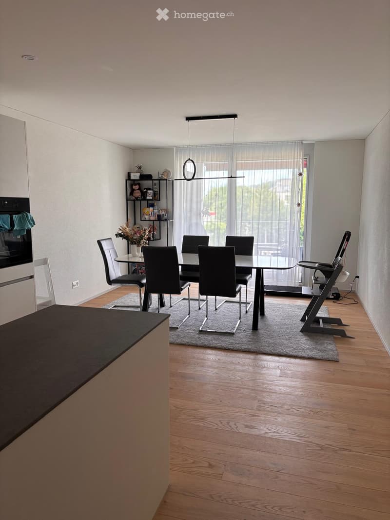 Modern dining area with a round glass table, six chairs, a baby chair, a shelf with decorative items, and a vase with flowers. The room features wooden flooring and large windows with sheer curtains.