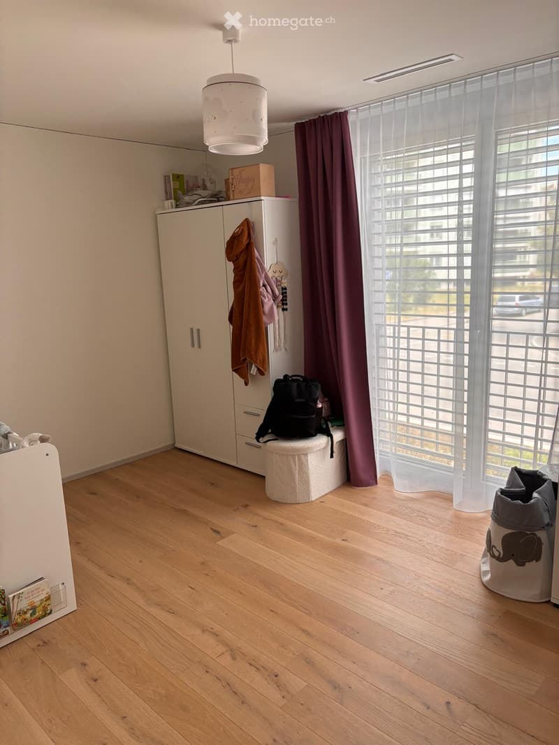 Bright room with wooden flooring, large window with curtains, white cabinet, stool, backpack, and decorative items.