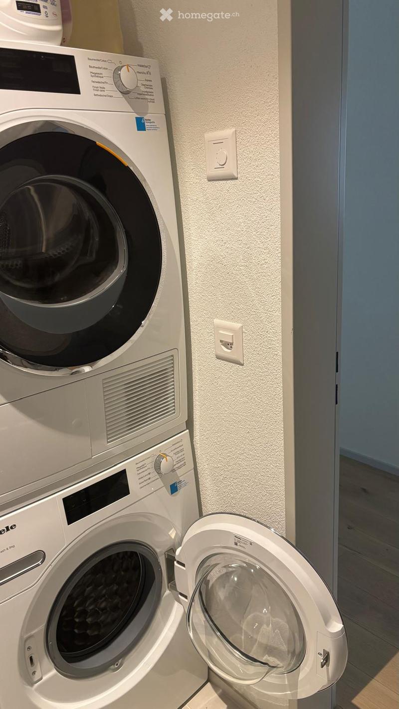 A modern bathroom with a white washing machine and dryer stacked vertically, featuring a control panel and a light switch on the wall.