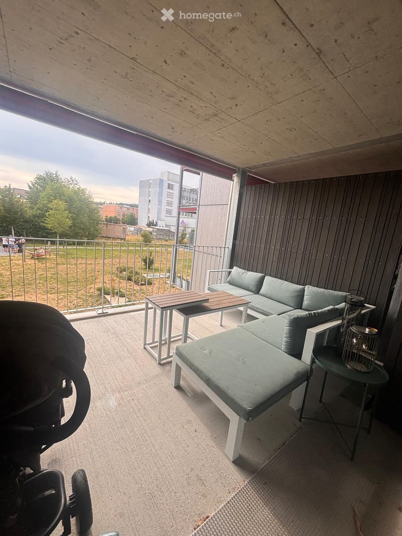 Modern balcony with grey sofa, ottoman, wooden table, side table, and lantern. Metal railing and concrete ceiling.
