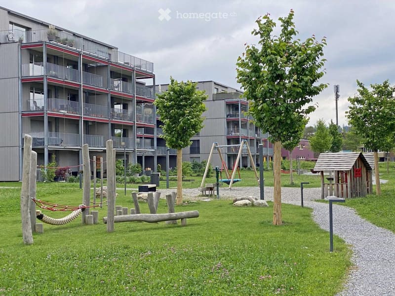 A modern residential complex with a well-maintained playground area featuring swings, a wooden playhouse, and a rope swing, surrounded by trees and grassy areas.