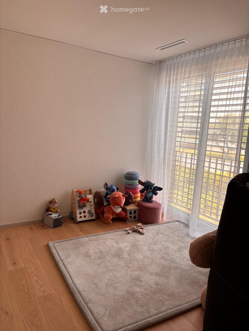 A cozy, well-lit room with wooden flooring, a large window with sheer curtains, and a variety of colorful toys including plush figures and a toy shelf.