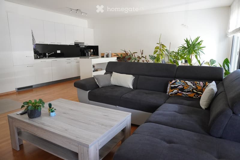 Modern living room with a grey sectional sofa, wooden coffee table, and kitchen area with white cabinets and black countertops.