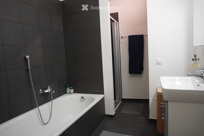 Modern bathroom with a white bathtub, chrome faucet, white sink, mirror, and dark towel hanging on a rack. The walls are tiled in dark gray, and the floor is black.