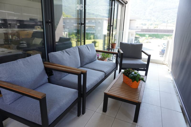 Balcony with grey sofa, armchair, small wooden table, and potted plants, overlooking a residential area.