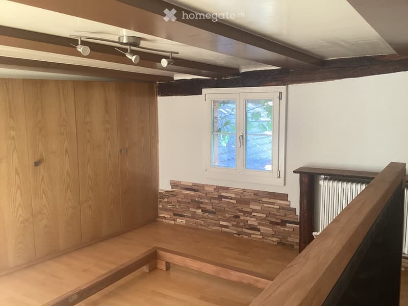 Empty room with wooden flooring, exposed wooden beams on the ceiling, a window with frosted glass, and a radiator under the window.