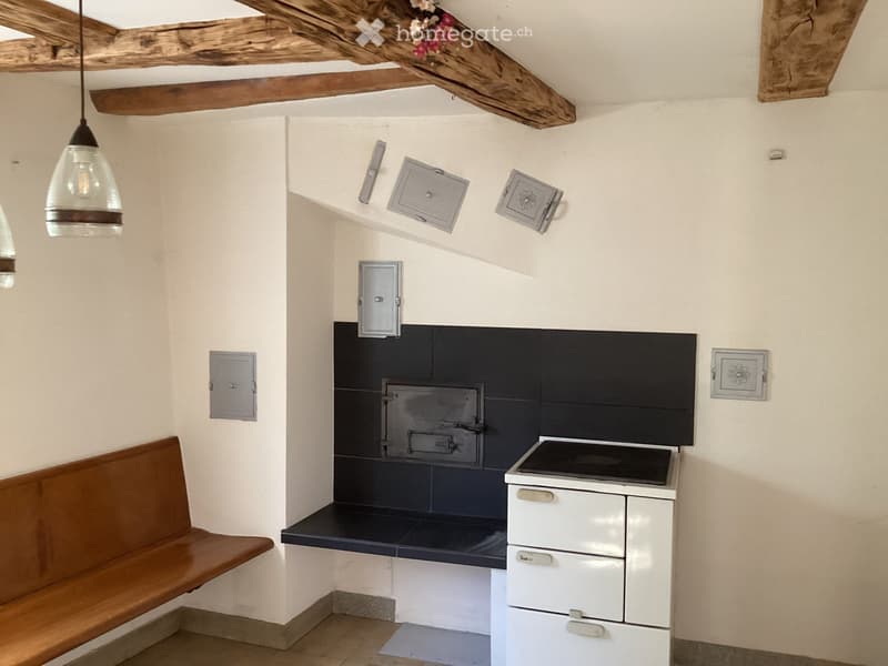 Cozy kitchen with exposed wooden beams, a wooden bench, a stove, and an oven. The walls are painted in a light beige color with dark blue tiles around the oven area.