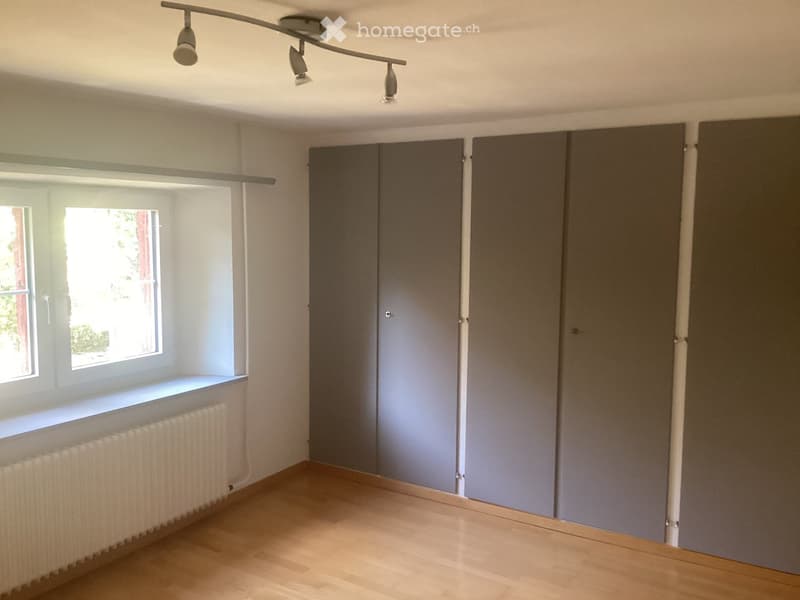 Empty room with wooden flooring, white walls, large window with white frame, radiator, and grey closet doors.