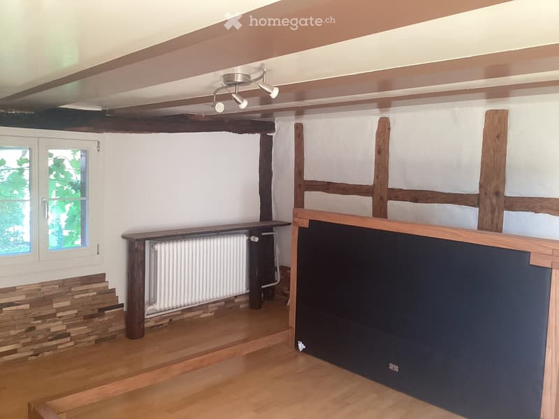 Empty room with wooden floor, exposed wooden beams, white walls, and a radiator. Features a stone accent wall and a window with a view of greenery.