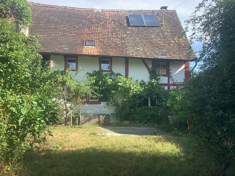 Two-story house with a tiled roof, white walls, and red wooden trim. The house has multiple windows and a small dormer window on the roof. The surrounding area includes a grassy lawn, trees, and a paved pathway leading to the house.