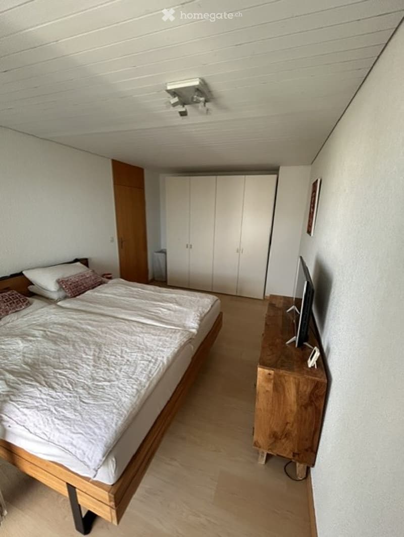 Modern bedroom with a wooden bed, white bedding, a nightstand with a screen, and a white wardrobe.