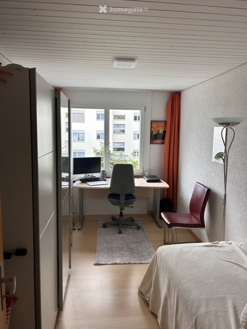 A modern, well-lit bedroom with a bed, desk, chair, computer, and a lamp. The room features wooden flooring, white walls, and a large window with a view of the city.