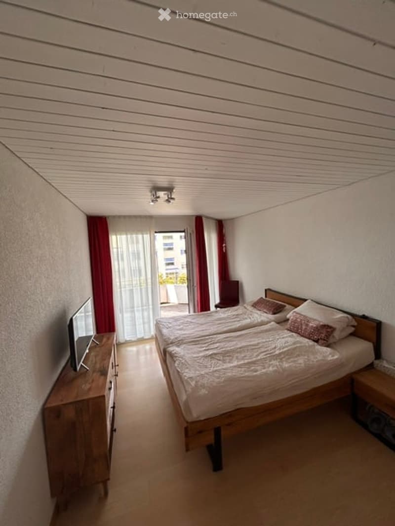Cozy bedroom with two single beds, a wooden nightstand, a chair, and a television. The room features red curtains and a large window with a view of the outside.
