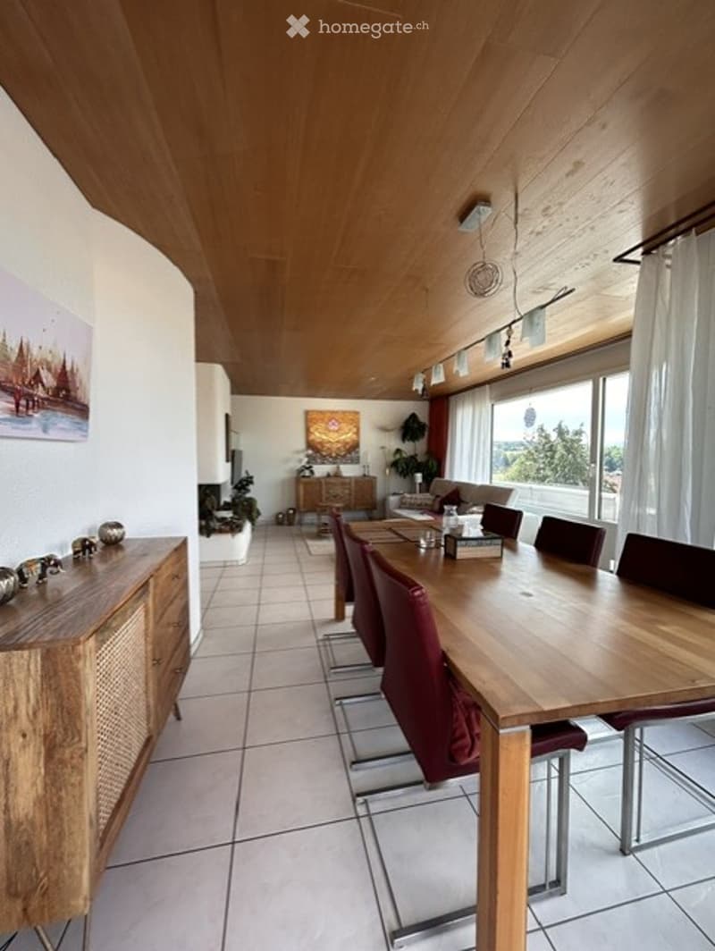 Bright dining area with a long wooden dining table surrounded by red chairs, a sofa in the background, and a wooden cabinet with decorative items. Large windows offer a scenic view.
