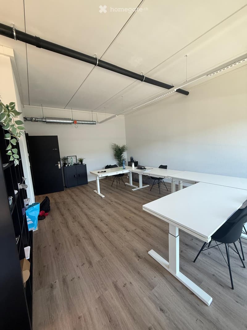 Modern office space with wooden flooring, white tables, black chairs, and a cabinet with plants.