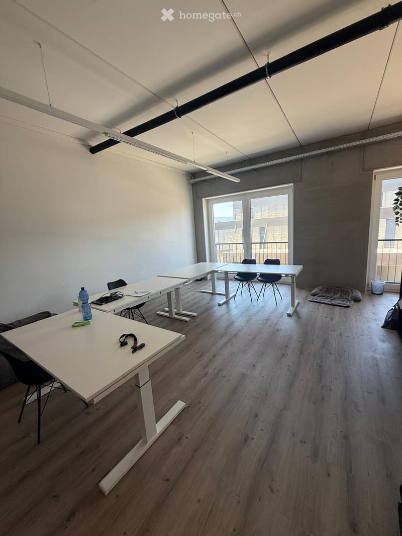 Bright, modern room with wooden flooring, white tables, and black chairs arranged in a collaborative workspace. Large windows and a balcony offer natural light and an outdoor view.