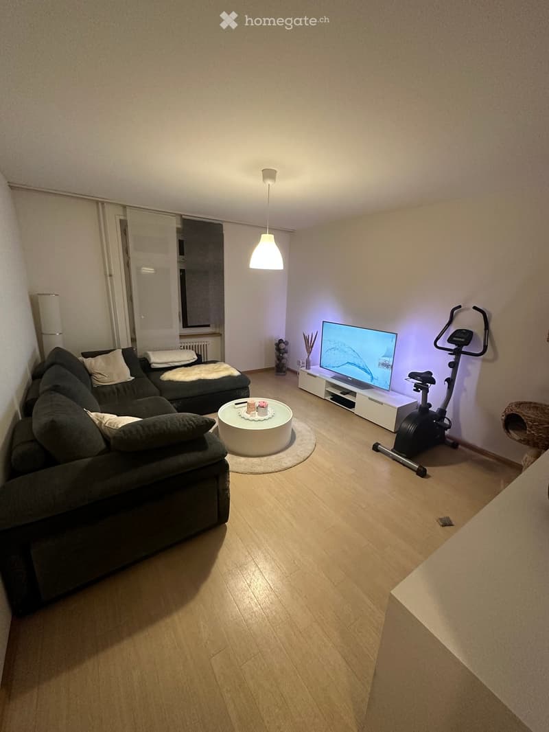 Living room with a dark grey sectional sofa, a round white coffee table, a flat-screen TV on a white stand, an exercise bike, and a cat tree. The room has light wooden flooring and white walls.