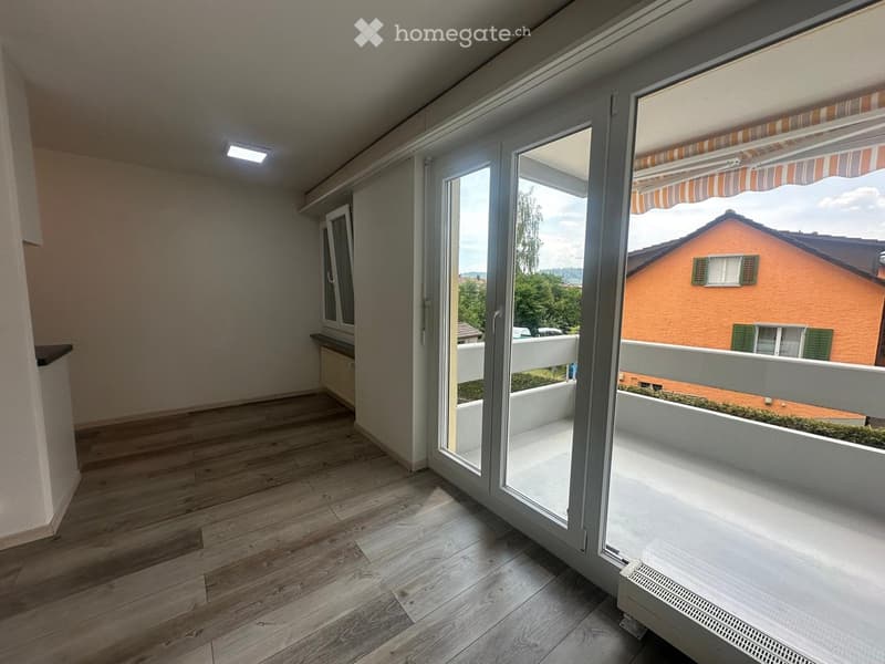 Empty room with wooden flooring, large sliding glass doors leading to a balcony, and a view of a neighboring house and greenery.