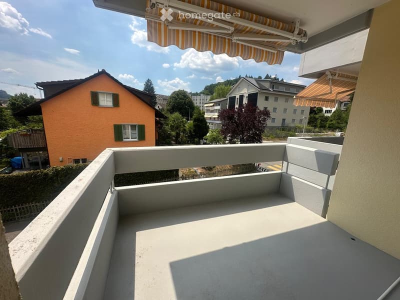 Balcony with white railing and striped awning, overlooking a residential area with houses and greenery.