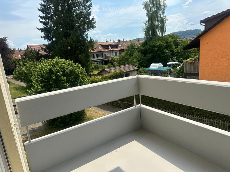 Balcony with white railings, overlooking a residential area with houses, trees, and a swimming pool in the distance.