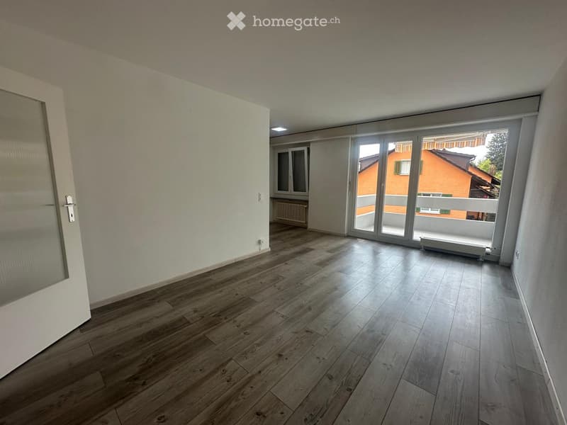 Empty room with wooden flooring, white walls, large sliding glass doors leading to a balcony, and a radiator visible in the background.