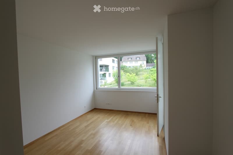 Empty room with light wooden flooring and white walls, featuring a large window with a view of neighboring buildings and greenery.