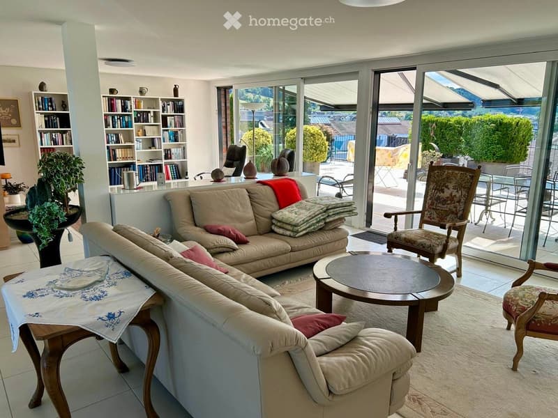 Bright living room with large windows and a view, featuring a beige sectional sofa, coffee table, wooden chairs, bookshelf, and decorative plants.