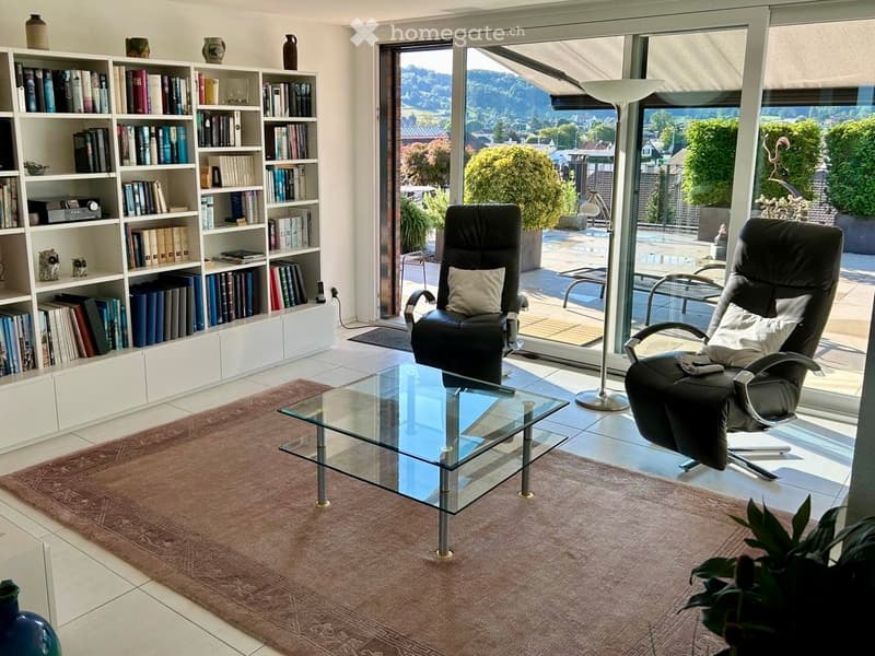 Modern living room with large windows offering a scenic view, featuring a glass coffee table, two black leather chairs, a bookshelf filled with books, and a floor lamp.