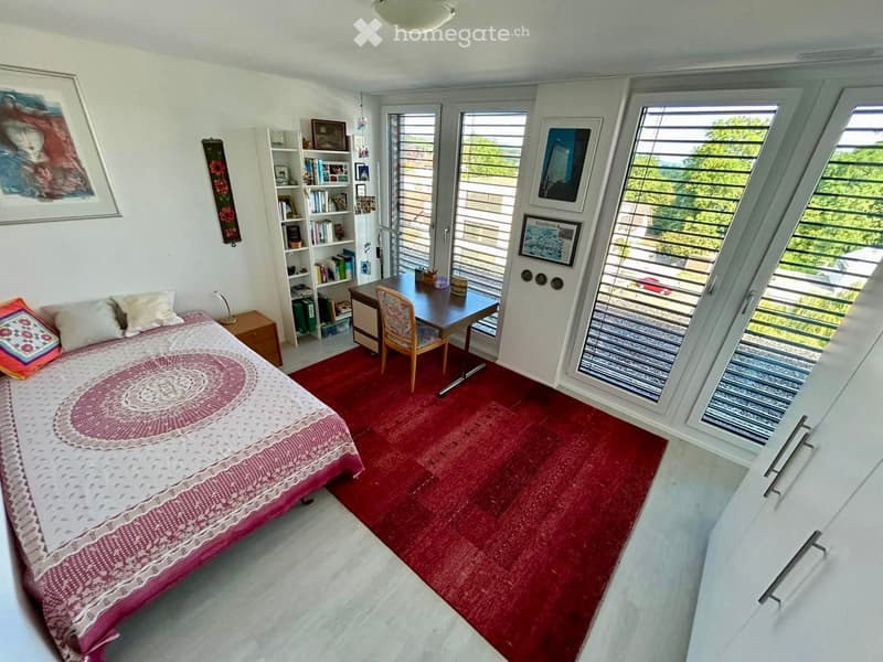 Bright bedroom with large windows and a balcony view, featuring a bed with a colorful bedspread, a wooden desk with a chair, a bookshelf, and a nightstand with a lamp.