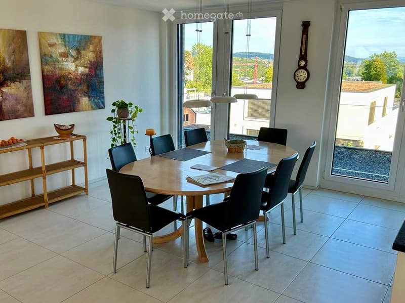 Bright dining room with a round wooden table surrounded by black chairs, a sideboard with a plant, and a large window offering a scenic view.