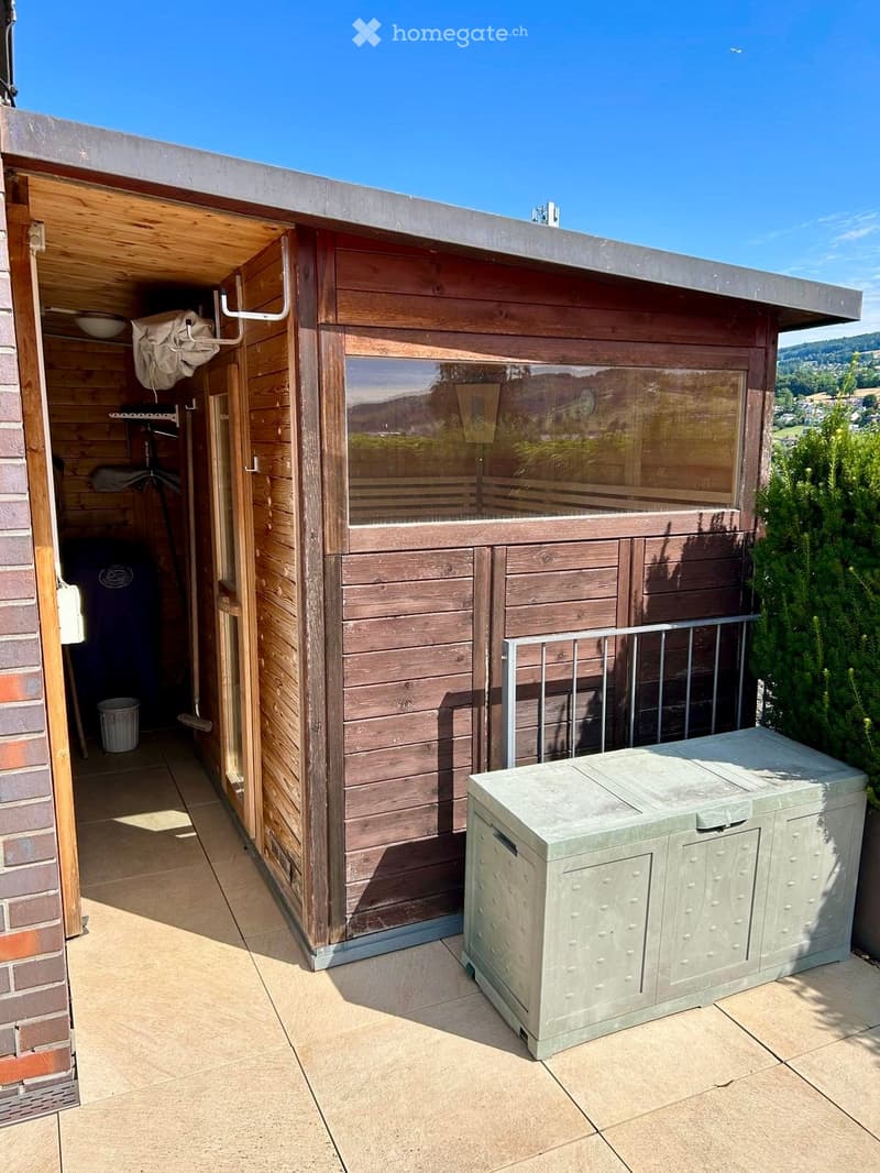 Wooden outdoor structure with large glass window, metal railing, and storage unit on tiled patio.