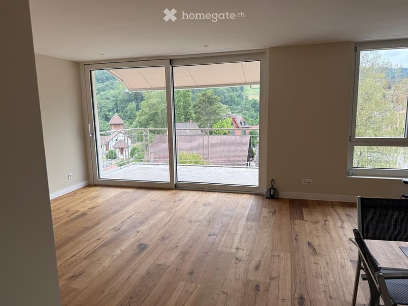 Empty room with wooden flooring, large sliding glass doors leading to a balcony with a scenic view of trees and houses.