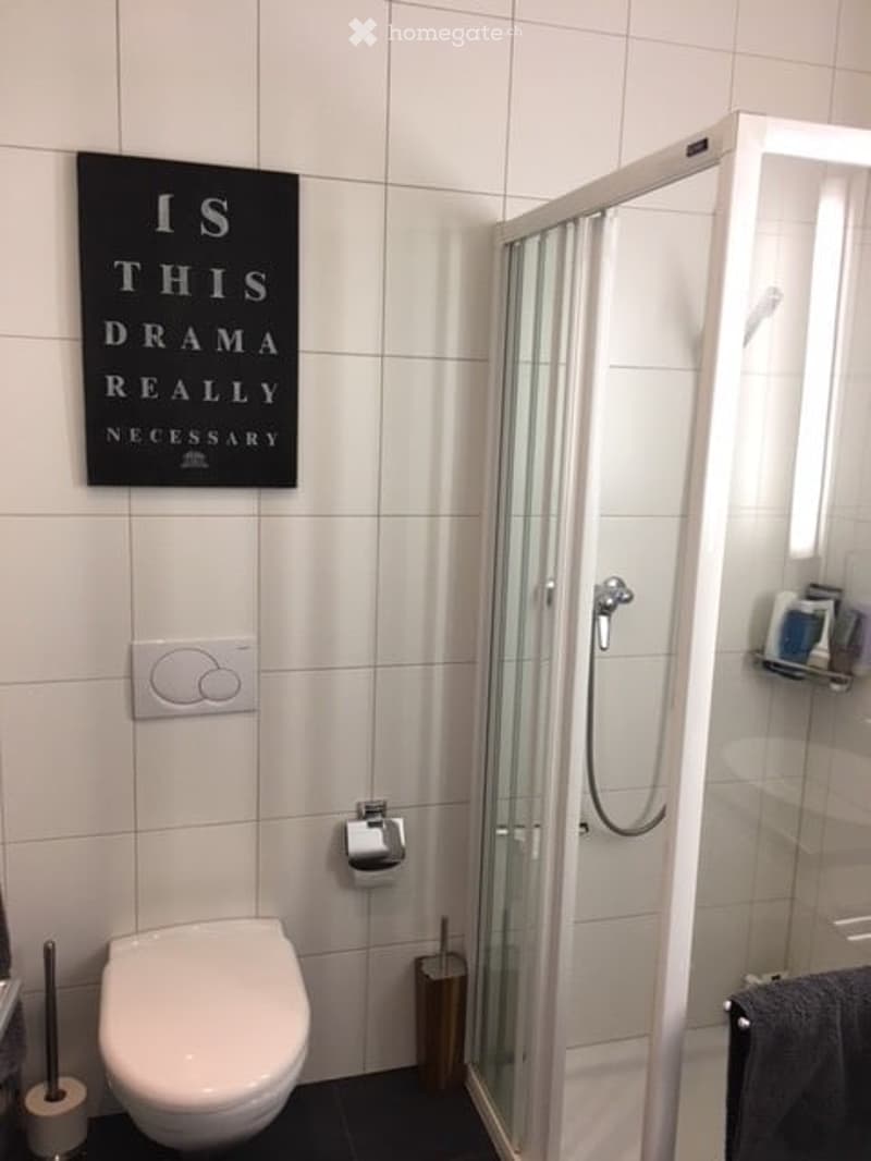 Modern bathroom with white tiles, a toilet, a glass-enclosed shower, a towel, and a decorative sign on the wall.