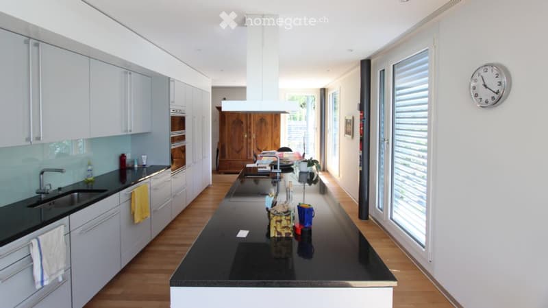 Modern kitchen with white cabinets, black countertop, stainless steel sink, and a central island. Features include a range hood, built-in oven, and large windows with blinds.