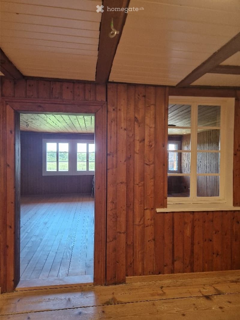 Empty room with wooden walls and floor, large windows, and an open doorway leading to another room.
