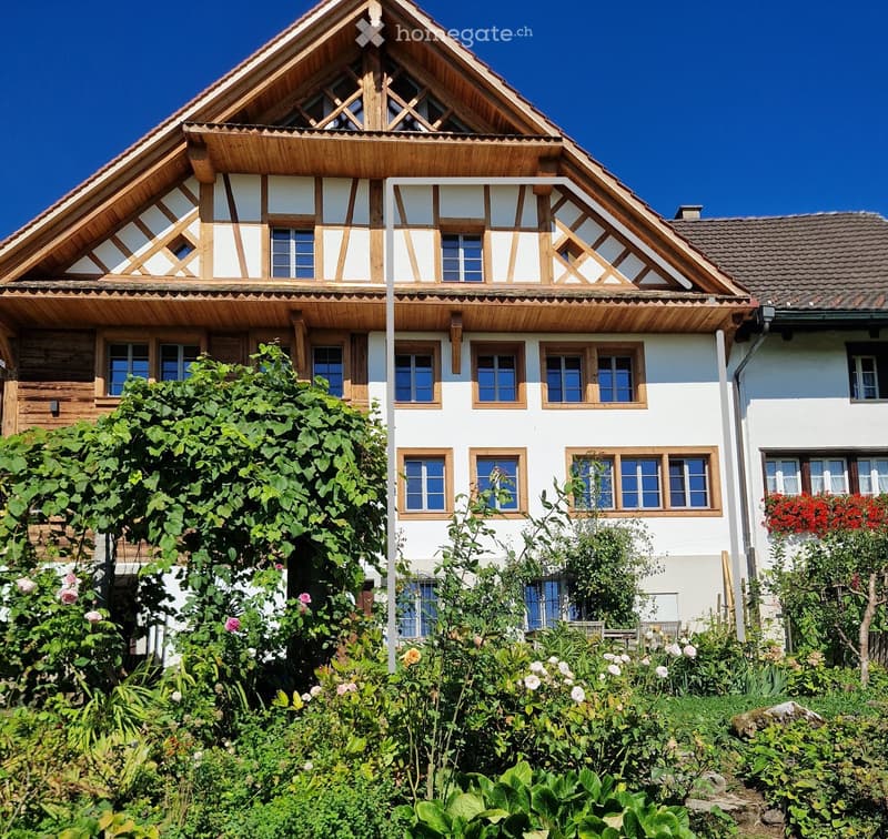 Two-story house with white walls, wooden trim, multiple windows, and a well-maintained garden with various plants and flowers.