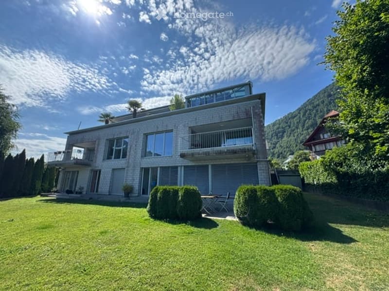 Modern two-story house with large windows, balconies, and a well-maintained lawn. The house features a contemporary design with clean lines and a mix of brick and glass.