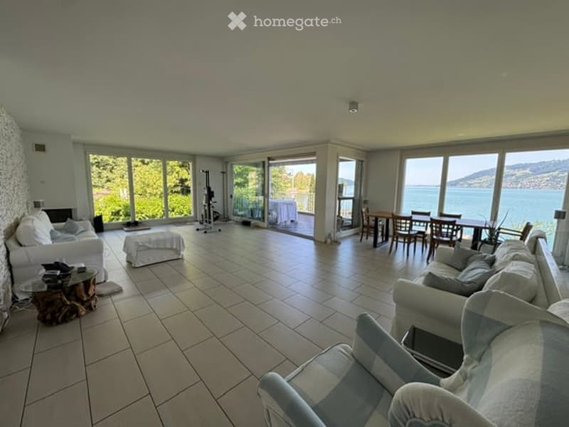 Bright, open living space with large windows offering a scenic view, featuring a sofa, ottoman, coffee table, dining area with a table and chairs, and a fireplace.