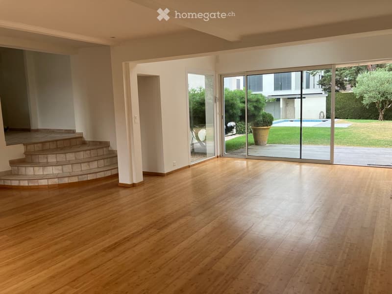 Empty room with wooden flooring, a staircase leading to an upper level, and large glass doors opening to a garden with a swimming pool.