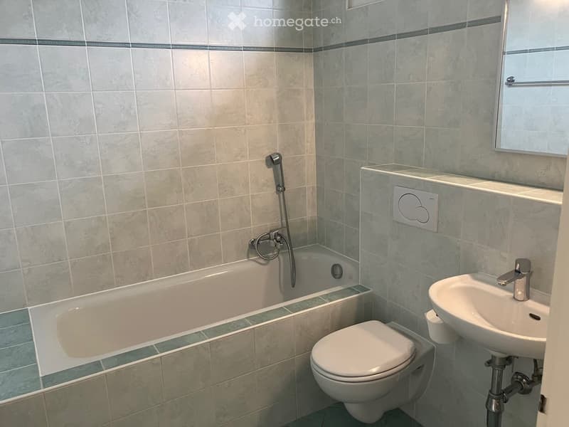 Modern bathroom with a bathtub, toilet, sink, and mirror. The walls are tiled with light grey tiles and a dark green border. A showerhead is attached to the wall above the bathtub.