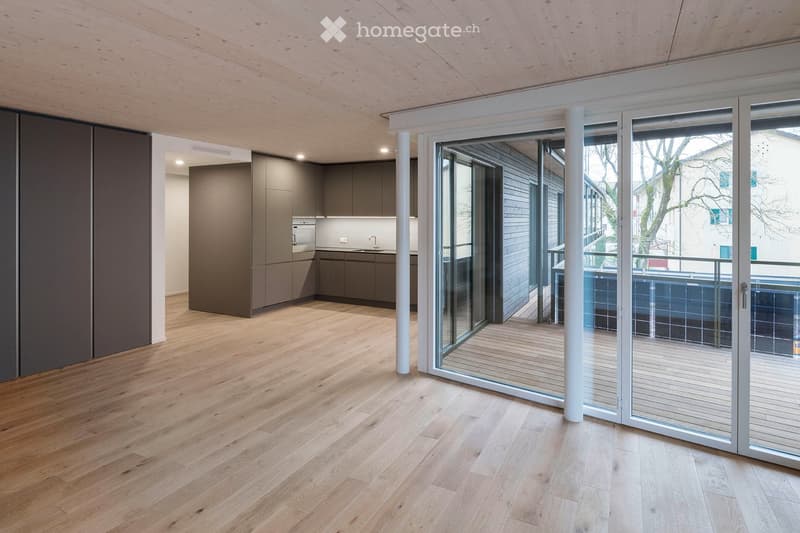 Open-plan living and kitchen area with light wooden flooring, modern grey kitchen cabinets, and a large glass sliding door leading to a balcony.