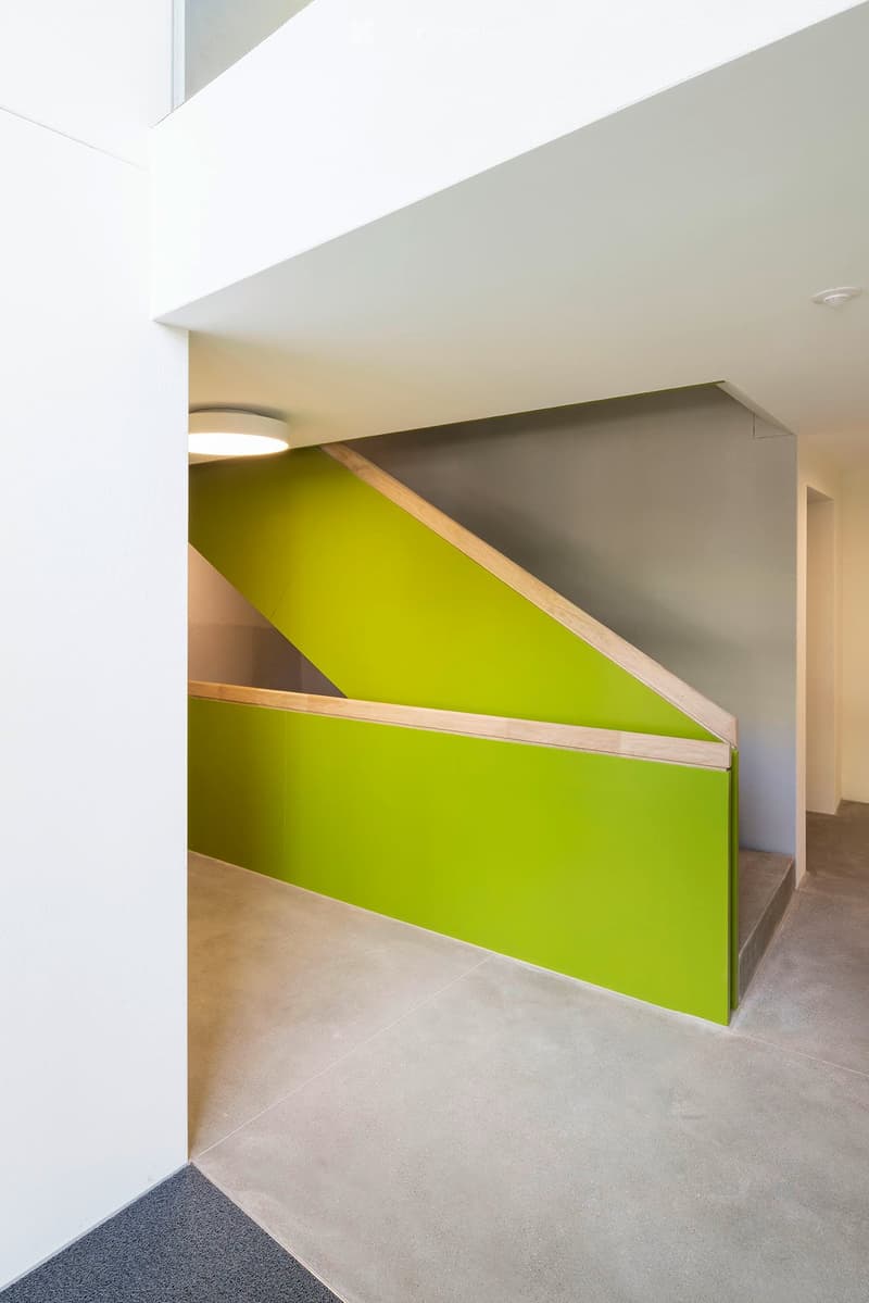 Modern hallway with a staircase featuring a green accent wall and wooden handrail, white walls, and grey tiled flooring.