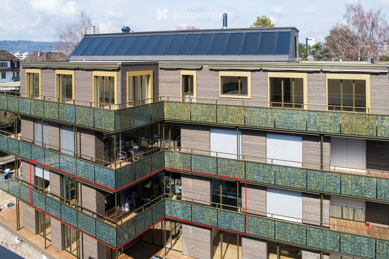 Modern multi-story residential building with balconies, large windows, and solar panels on the roof. The facade features a combination of wood and stone textures, with multiple balconies and outdoor seating areas visible.