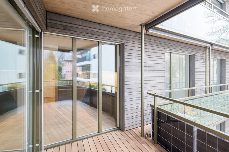 Modern balcony with wooden decking, large glass sliding doors, and a sleek metal railing.