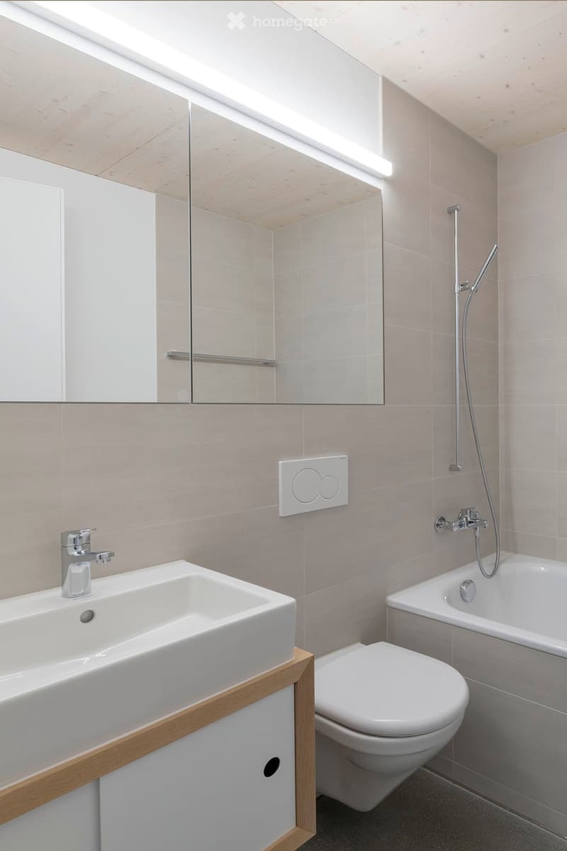 Modern bathroom with a white sink, toilet, bathtub, and showerhead. Features a large mirror with built-in lighting and sleek, contemporary design.