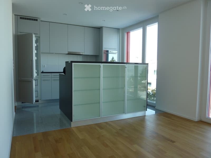 Modern kitchen with light grey cabinets, a central island with glass panels, and a refrigerator. The floor is wooden, and there are large windows letting in natural light.