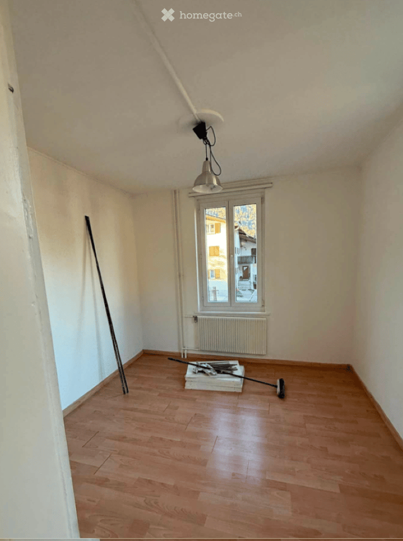 Empty room with wooden flooring, white walls, a single window with a radiator below, and a broom leaning against the wall.