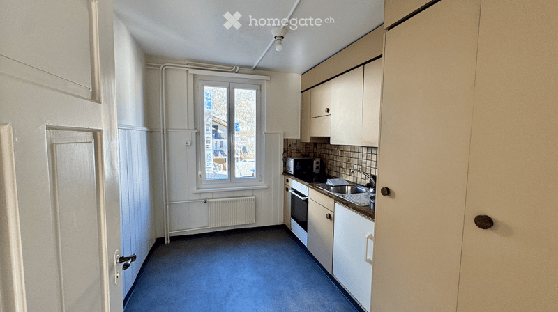 Compact kitchen with white cabinets, a sink, microwave, and oven. The space features a window with a view of the outside and a radiator below it.